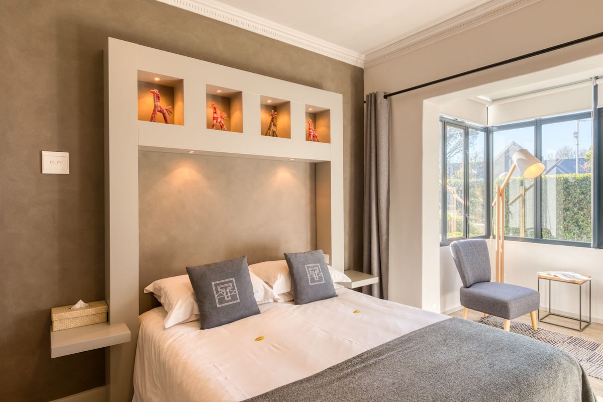 A modern hotel room with a large bed, a desk, and a chair. There are four illuminated shelves above the bed displaying decorative items.