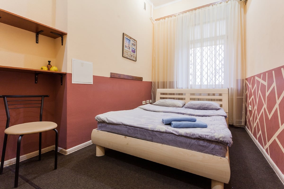 A single bed with a wooden frame, a chair, a small table with a vase, and a window with sheer curtains.