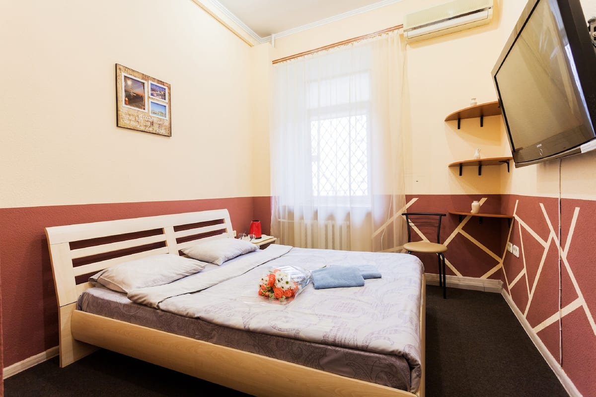 A neatly made bed with a floral arrangement, a bedside table with a red lamp, and a TV mounted on the wall.