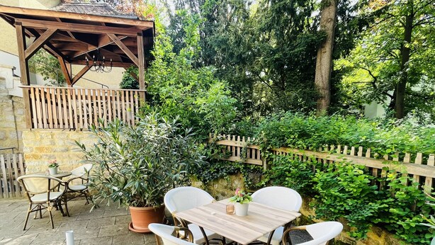 An outdoor patio with a wooden table and wicker chairs, surrounded by lush greenery and a stone wall. - Apartments at the Salvatorkirche - Apartment II (Ketschendorf)