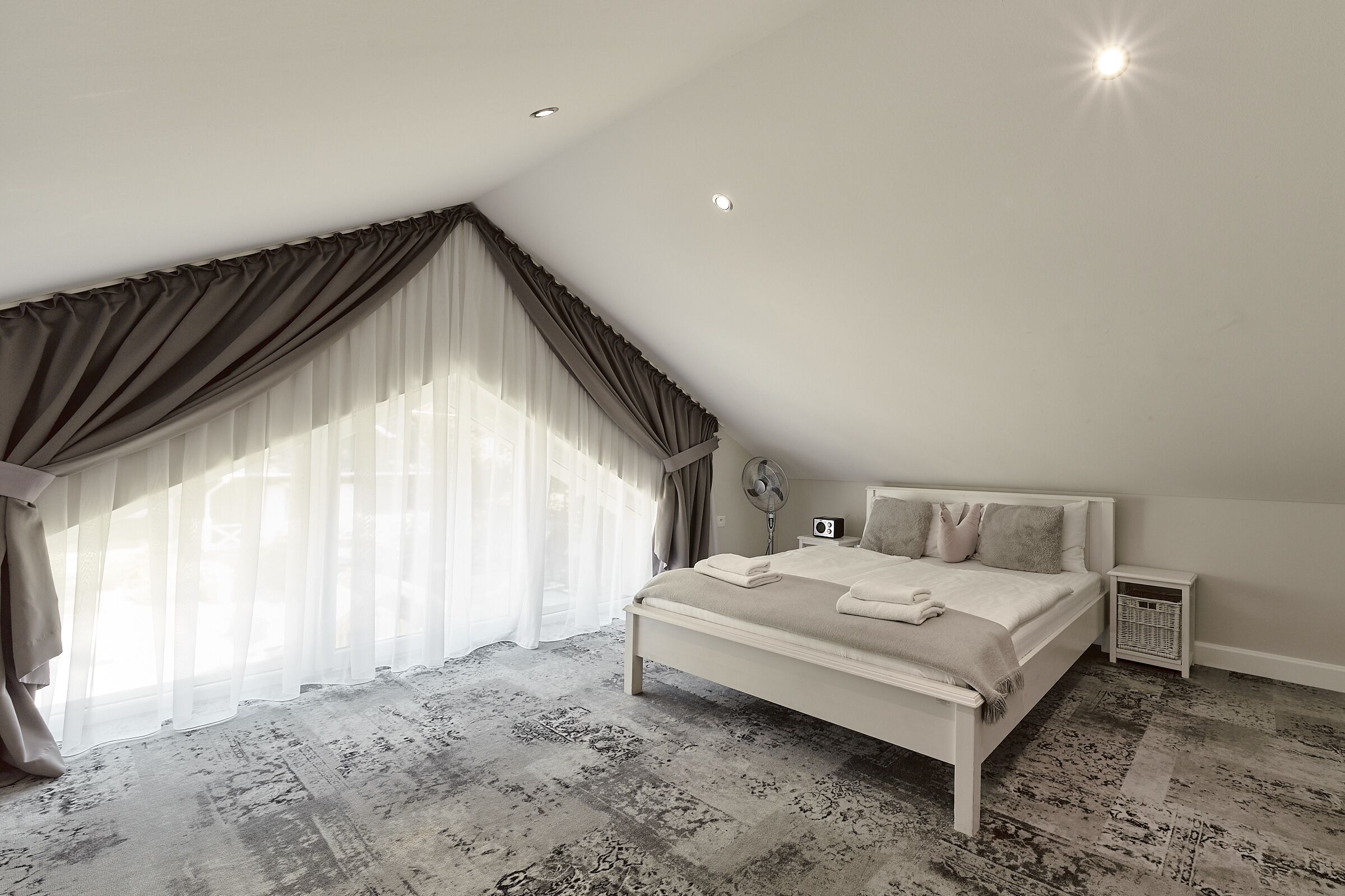 Un dormitorio con una cama grande, un ventilador de techo, una mesita de noche y una ventana con cortinas.