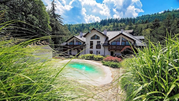 Une maison avec une piscine, entourée de verdure et située au pied d’une colline boisée.
