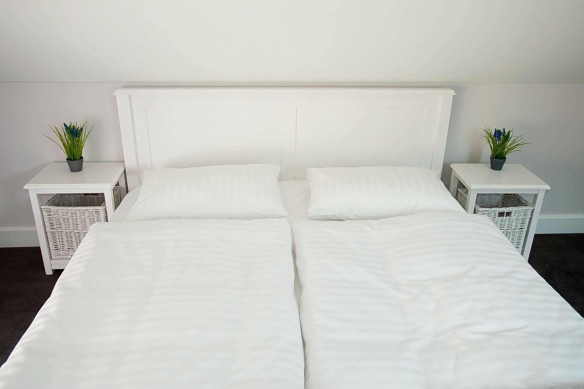 A double bed with white bedding, two nightstands with wicker baskets, and small potted plants.