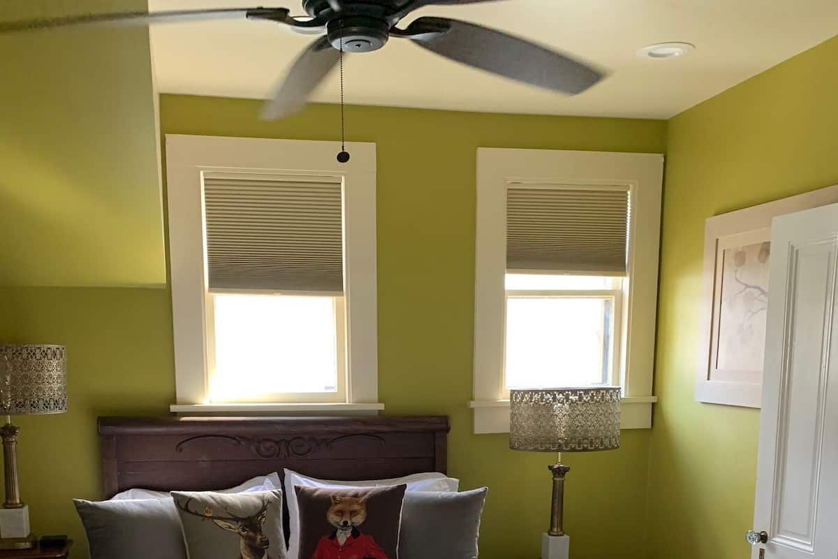 A bedroom with a bed, two windows with blinds, a ceiling fan, and a door.