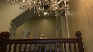 A chandelier with multiple crystal arms and lights, a wooden railing, and a staircase in the background. - The Hygrade Inn (Emporium)