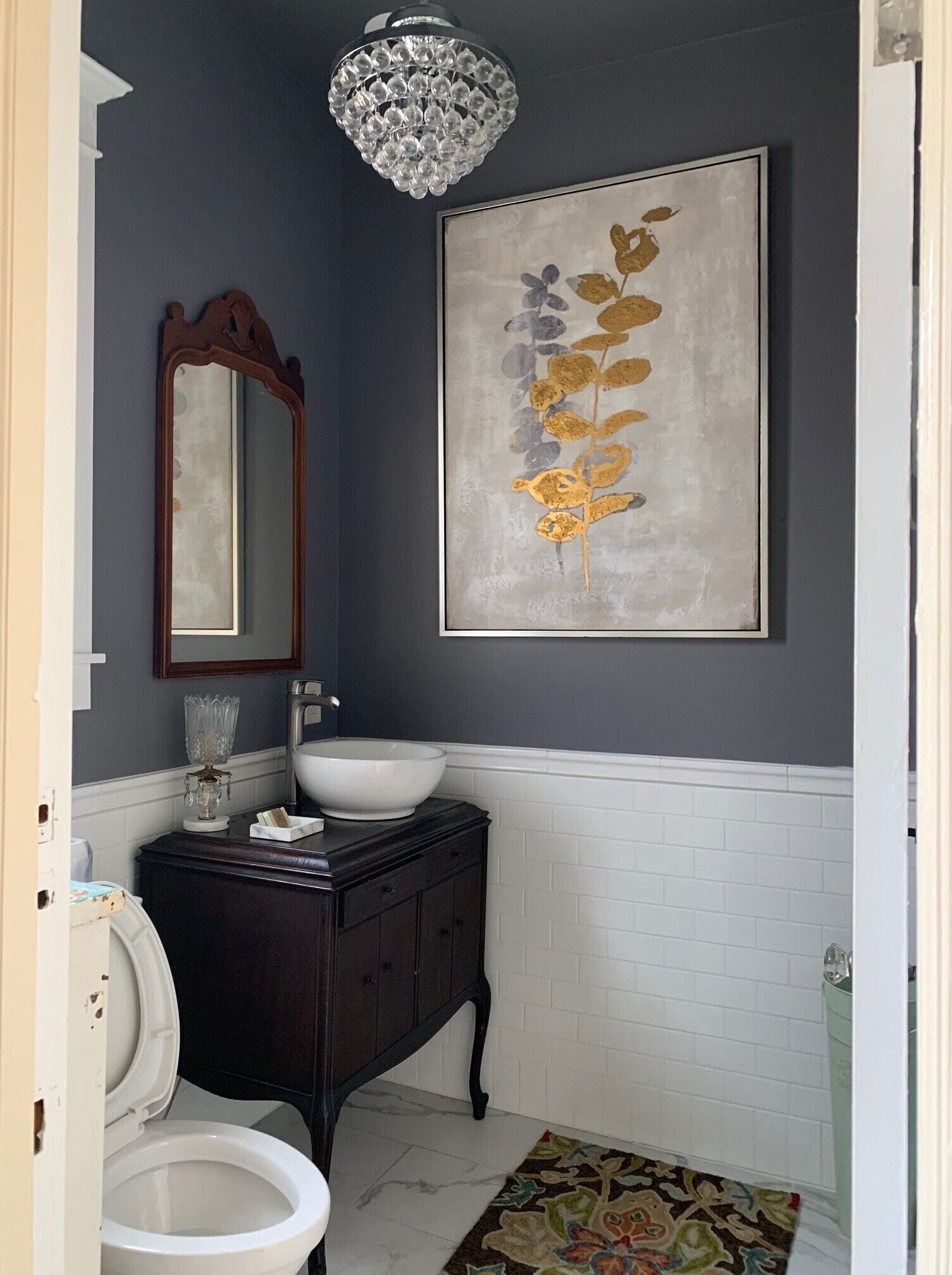 A bathroom with a dark wall, a white sink, a wooden vanity, a large mirror, and a chandelier.