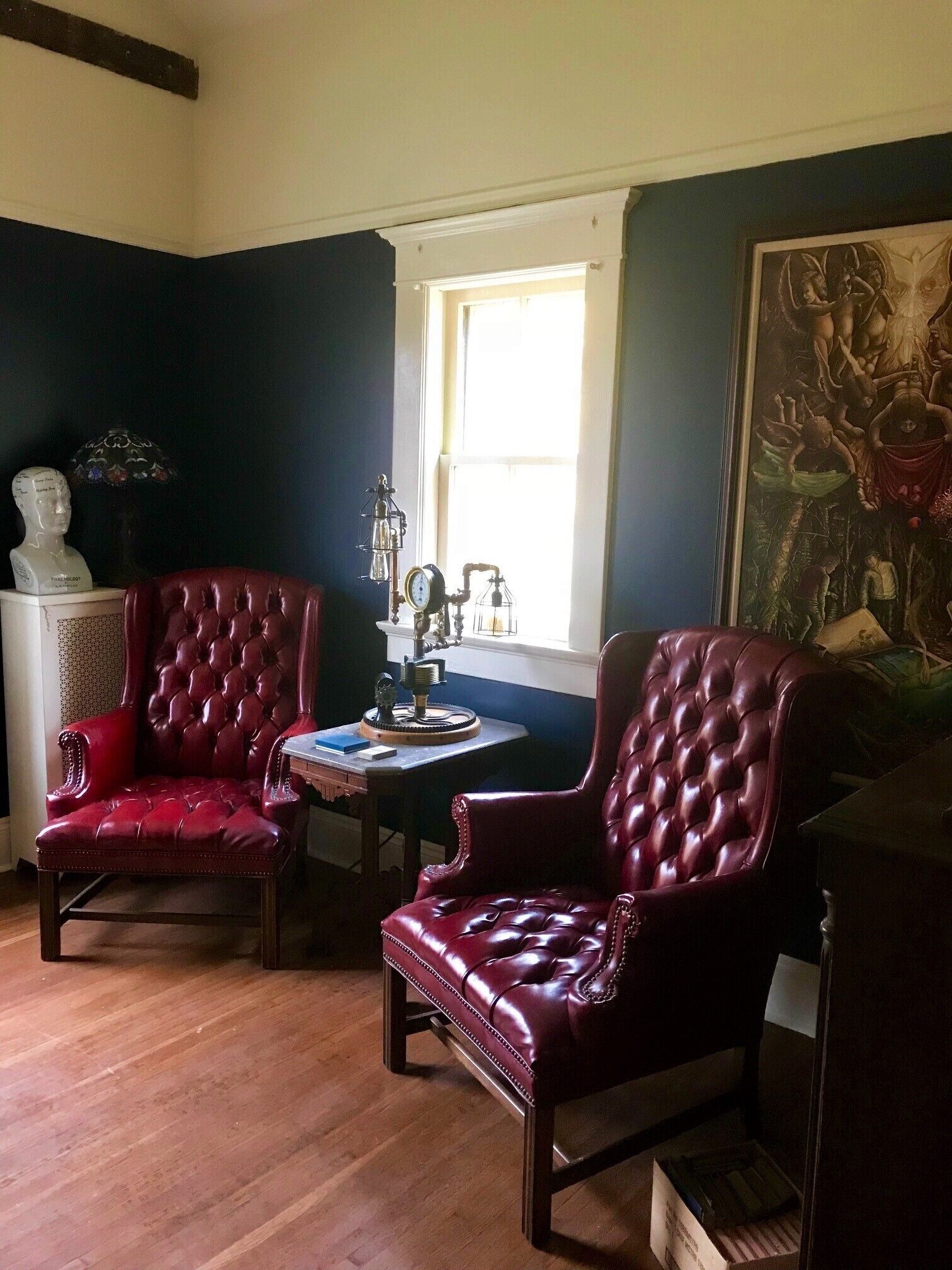 Una habitación con dos sillones de cuero rojo, una mesita con un reloj, un busto sobre un pedestal y un cuadro grande en la pared.