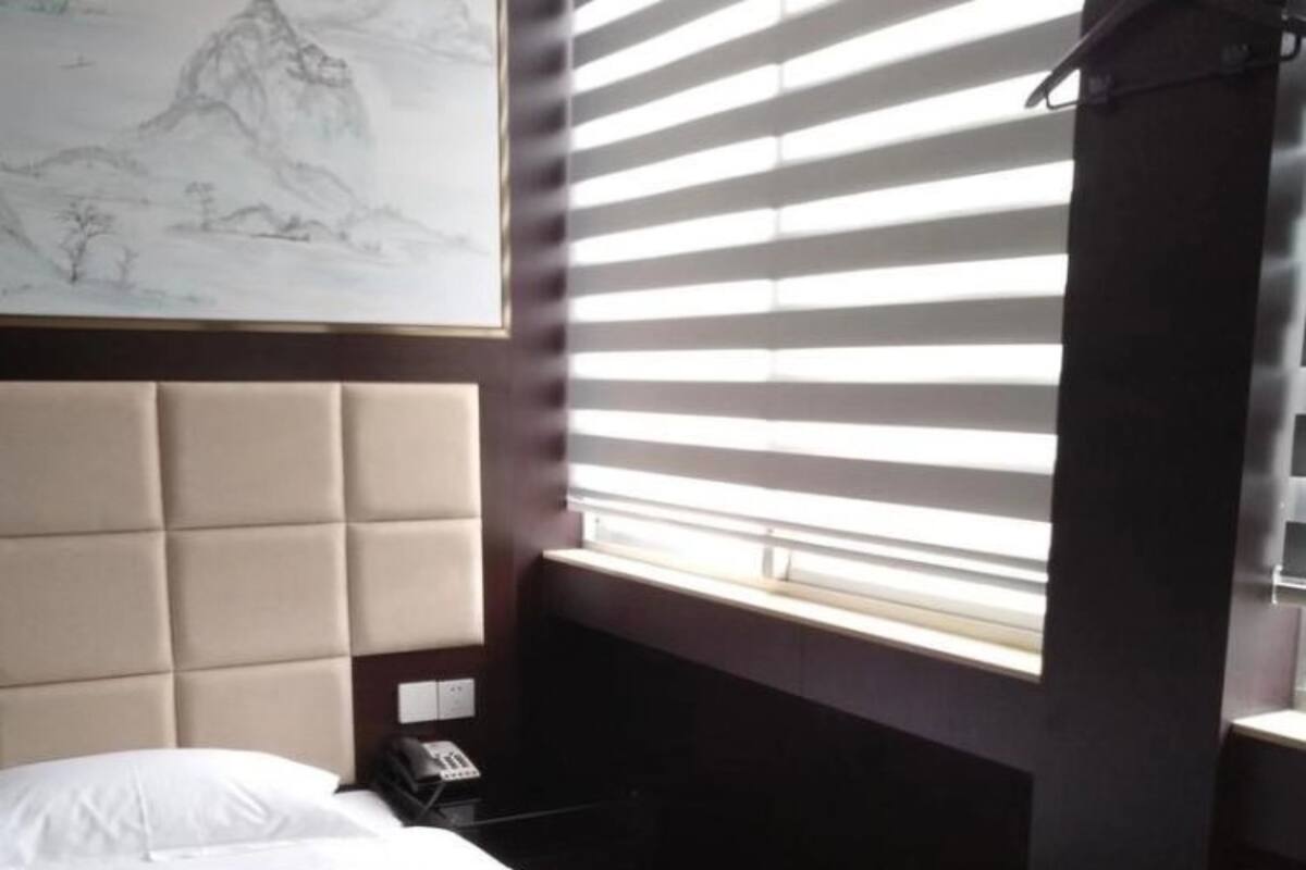 Hotelkamer met een bed, hoofdeinde, raam met jaloezieën, telefoon en een kunstwerk aan de muur.