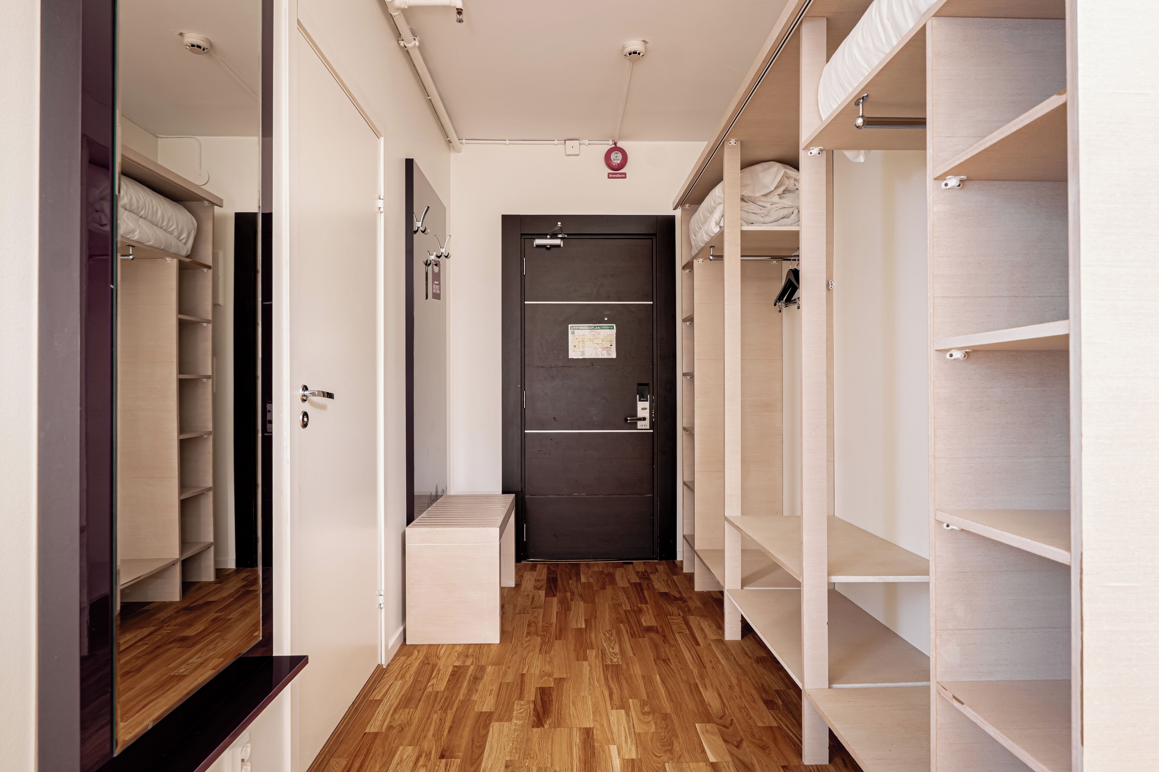 Un couloir d’hôtel avec un plancher en bois, une porte sombre et des étagères blanches de part et d’autre.