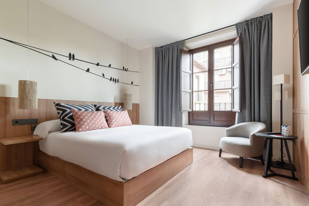 A modern hotel room with a large bed, a small armchair, a side table, and a window with curtains.