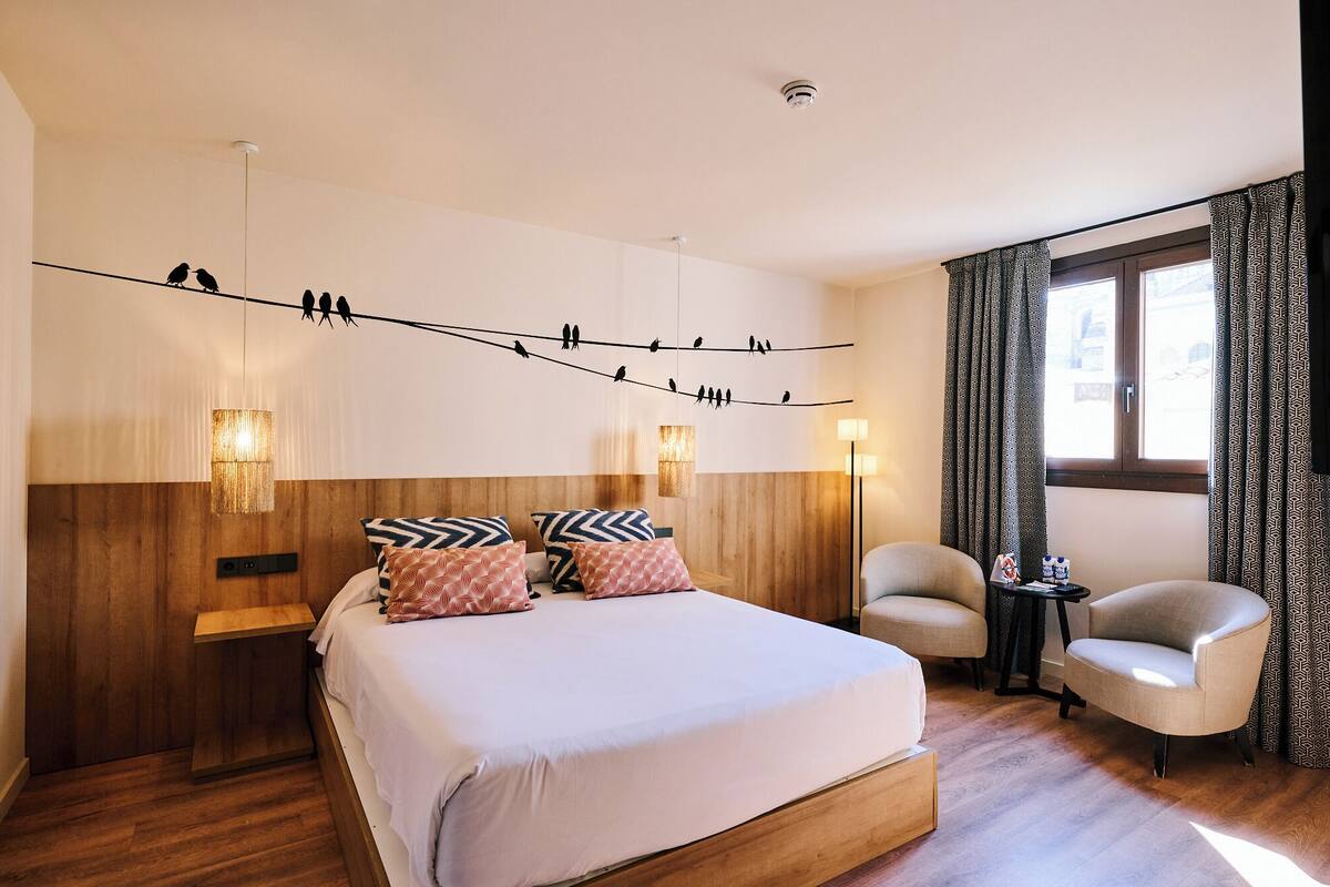 A hotel room with a wooden headboard, a large bed with patterned pillows, two bedside lamps, a small table with a chair, and a window with patterned curtains.