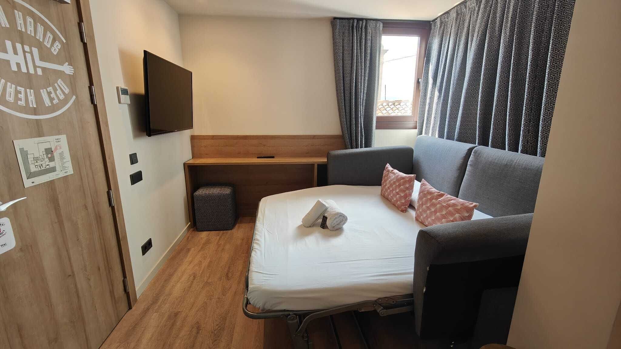 Habitació d'hotel senzilla amb un llit, escriptori, televisió penjada a la paret i una finestra amb cortines estampades.