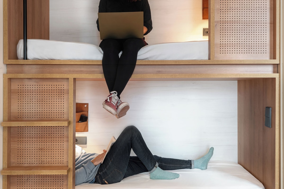A bunk bed with a person sitting on the top bunk using a laptop. The room has wooden walls and a white mattress.