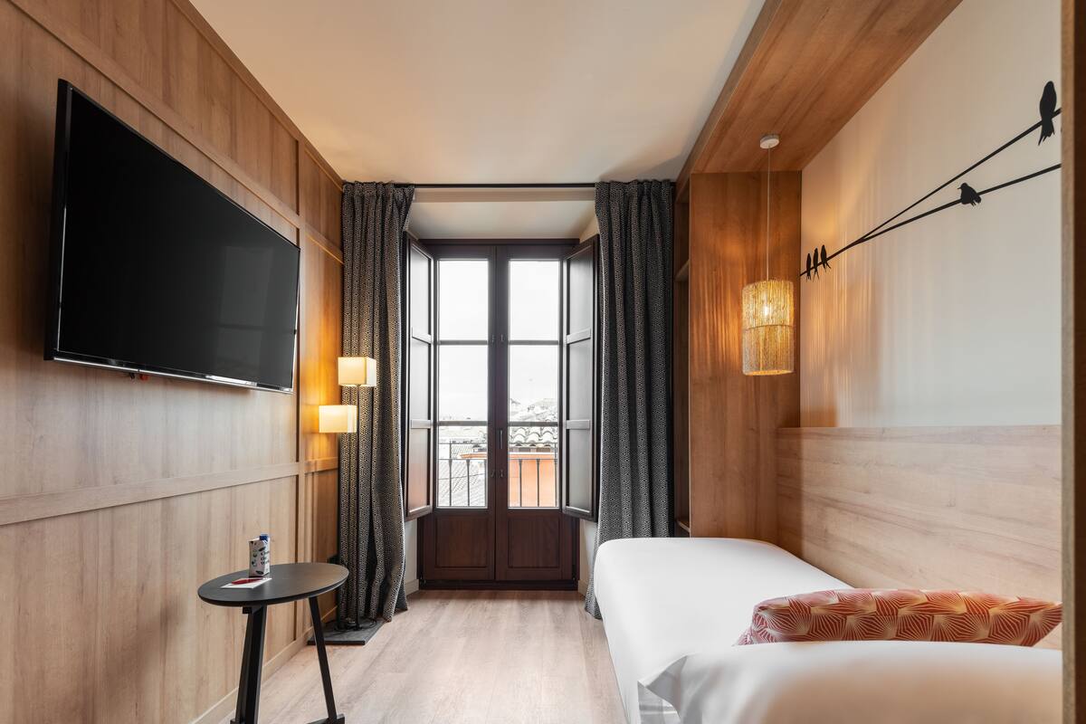 A hotel room with a bed, a TV mounted on the wall, a small table with a bottle, and a window with curtains.