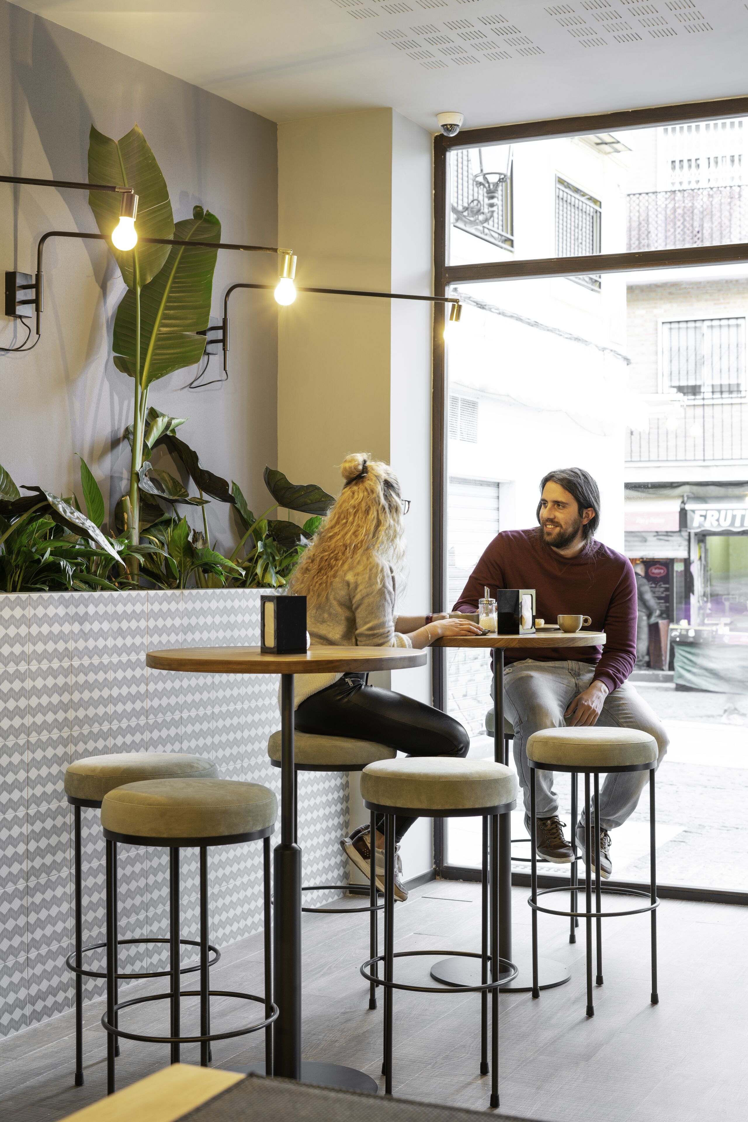 Um café moderno com uma planta grande, um cão sentado a uma mesa e uma pessoa sentada em frente ao cão.