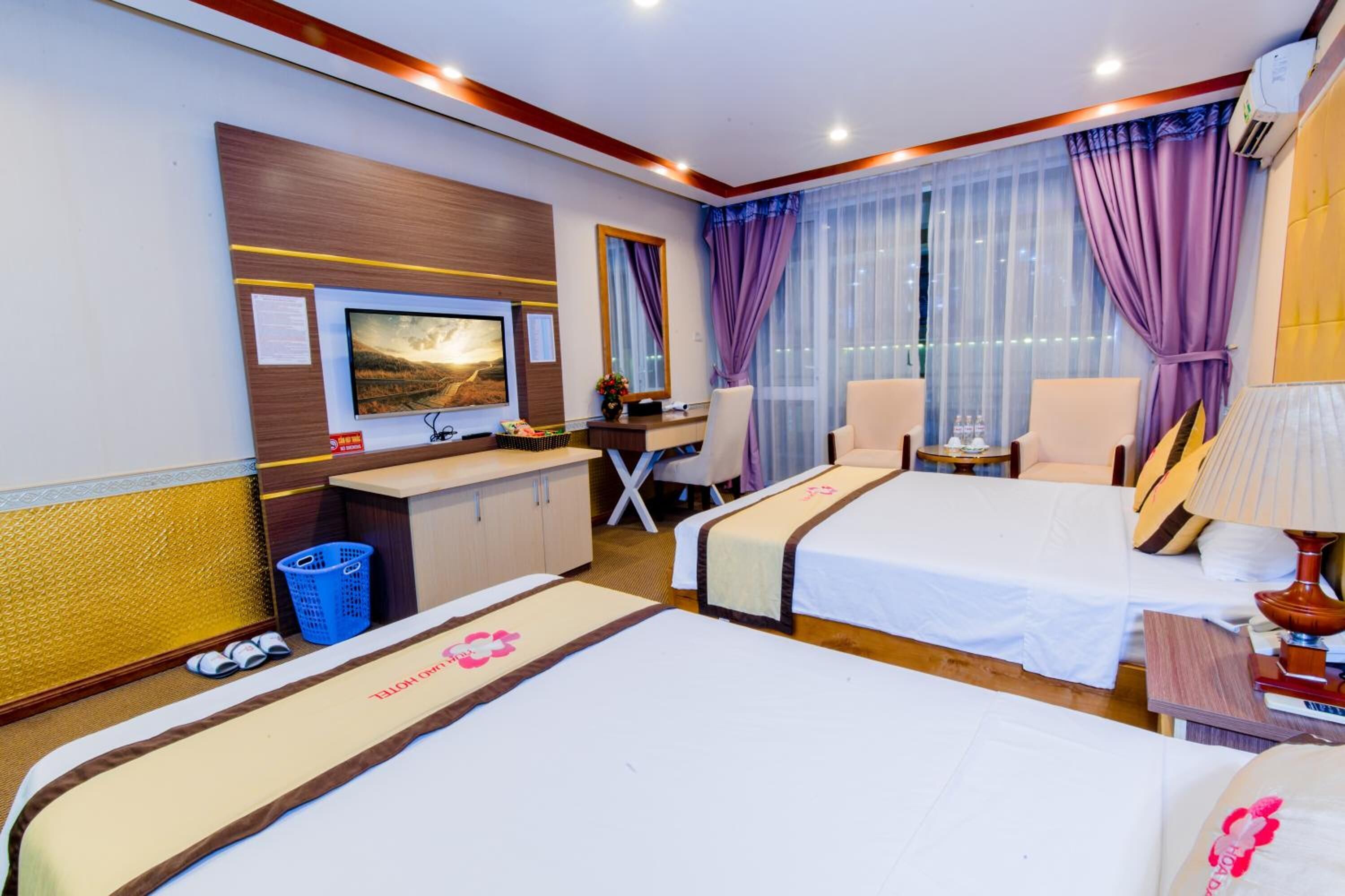 Quarto de hotel com duas camas, uma escrivaninha, uma cadeira, televisão e uma janela grande com cortinas.
