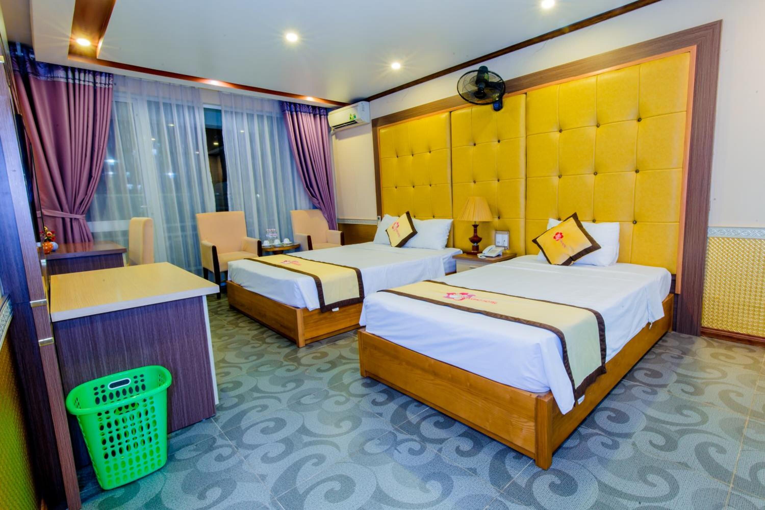 A hotel room with two beds, a desk, a chair, a green basket, and a window with curtains.