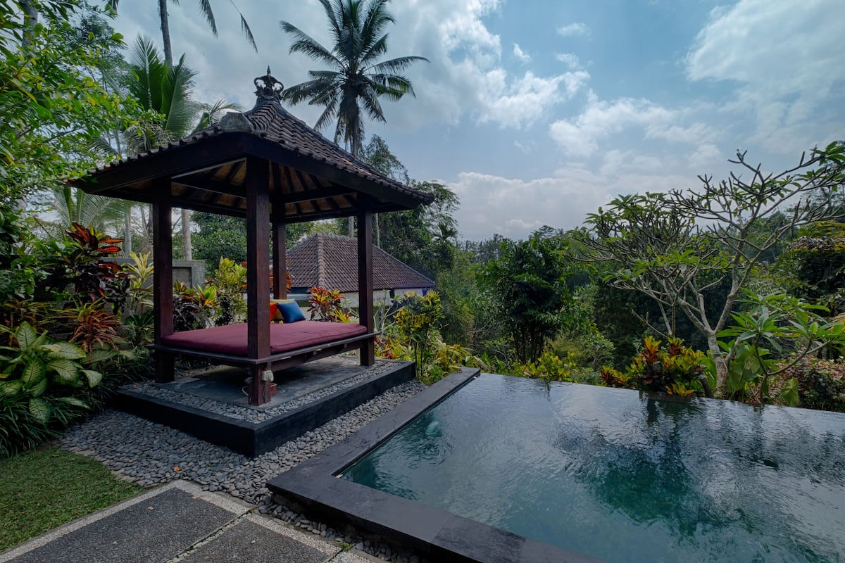A gazebo with a red cushioned bench, surrounded by lush greenery and a clear pool.