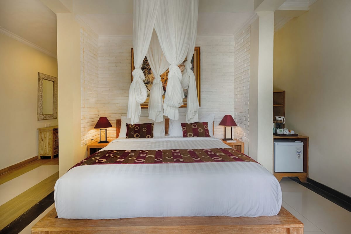 A large bed with a canopy, two bedside tables with lamps, a small refrigerator, and a mirror on the wall.