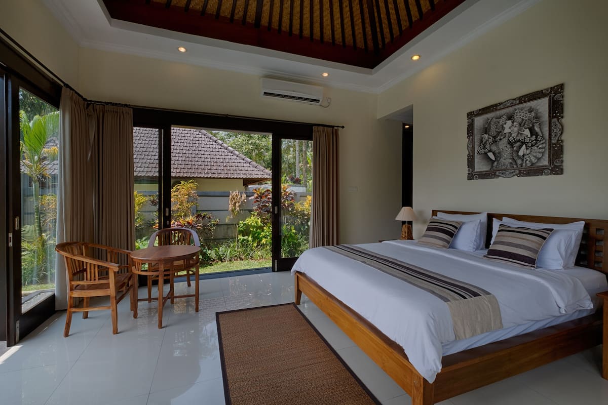 A bedroom with a large bed, a wooden chair, a table, and a view of a garden through a sliding door.