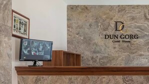 A reception desk with a monitor, a stone wall with the logo "DUN GORG Guest House", and a wooden counter. - Dun Gorg Guest House (Marsaxlokk)
