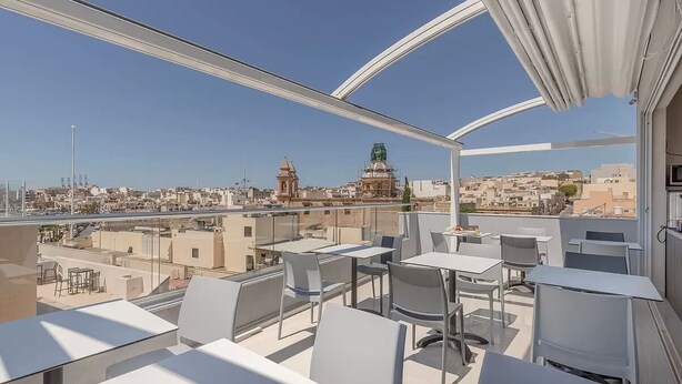 A rooftop terrace with tables and chairs, offering a view of a cityscape including a prominent dome and buildings. - Dun Gorg Guest House (Marsaxlokk)