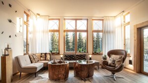 Un salon chaleureux avec un canapé, deux petites tables basses en bois et un fauteuil en cuir. De grandes fenêtres offrent une vue sur les arbres et une terrasse en bois.
