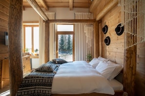 A wooden cabin bedroom with a bed, a dresser, and a window overlooking a snowy landscape.