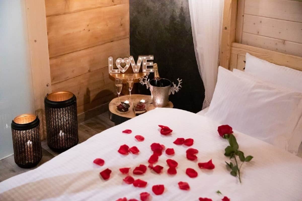 Een net opgemaakt bed met rozenblaadjes en een enkele roos, een kandelaar met een kaars, en een decoratief bordje met de tekst 'LOVE' in een kamer met houten wanden en een gordijn.