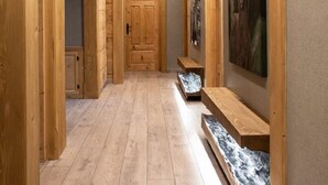 Un couloir en bois, avec une porte en bois au fond, des bancs le long des murs et un tableau sur le mur de droite.