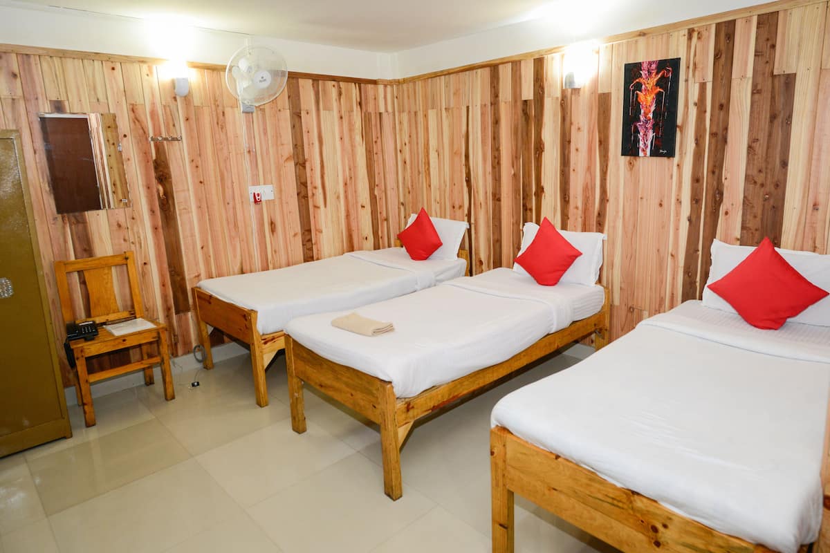 A room with three wooden beds, each with white bedding and red pillows. There is a wooden chair and a small table with a fan on the wall.