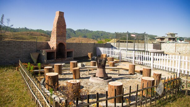 A rustic outdoor area with a brick oven, wooden stumps, and a white fence.