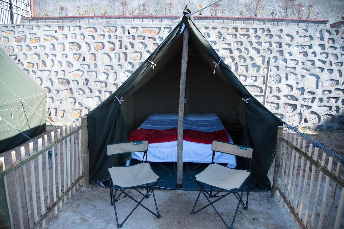 A tent with a bed and two chairs inside, set against a stone wall.