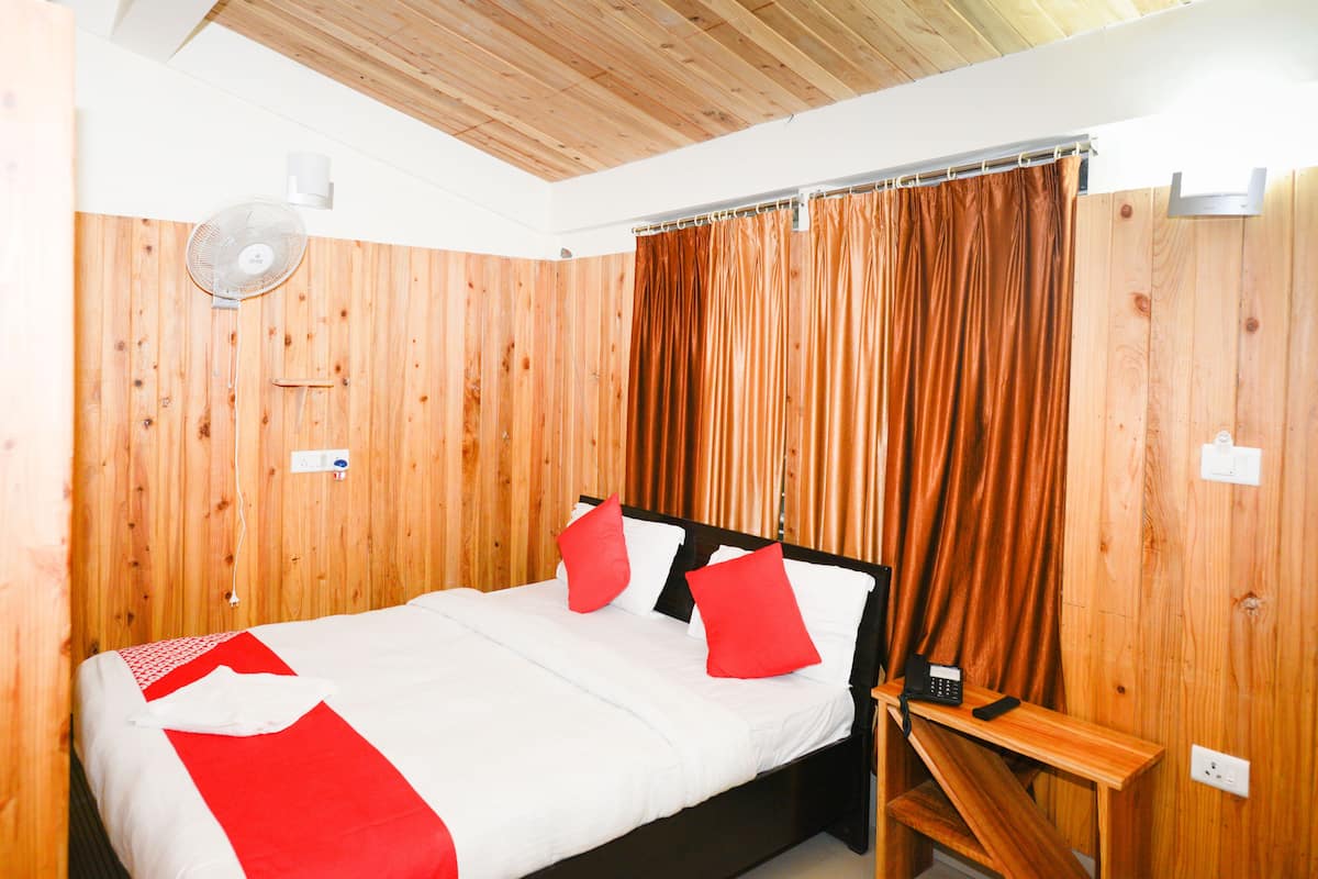 A room with a wooden paneled wall, a bed with white bedding and red pillows, a bedside table with a phone, and a ceiling fan.