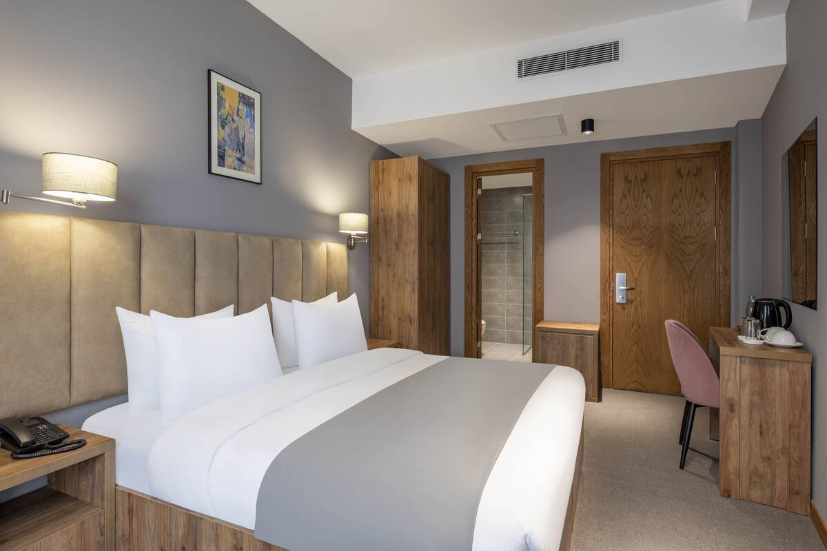 Una habitación de hotel moderna con una cama grande, lámparas de noche, un armario de madera, un escritorio con una silla y un área de ducha.