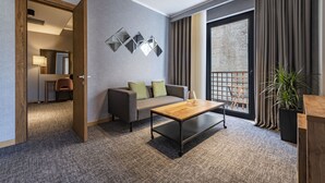 A modern hotel room with a grey carpet, a wooden coffee table, a grey sofa with green cushions, a large window with curtains, and a potted plant. - BARNOVI BOUTIQUE HOTEL (Tbilisi)