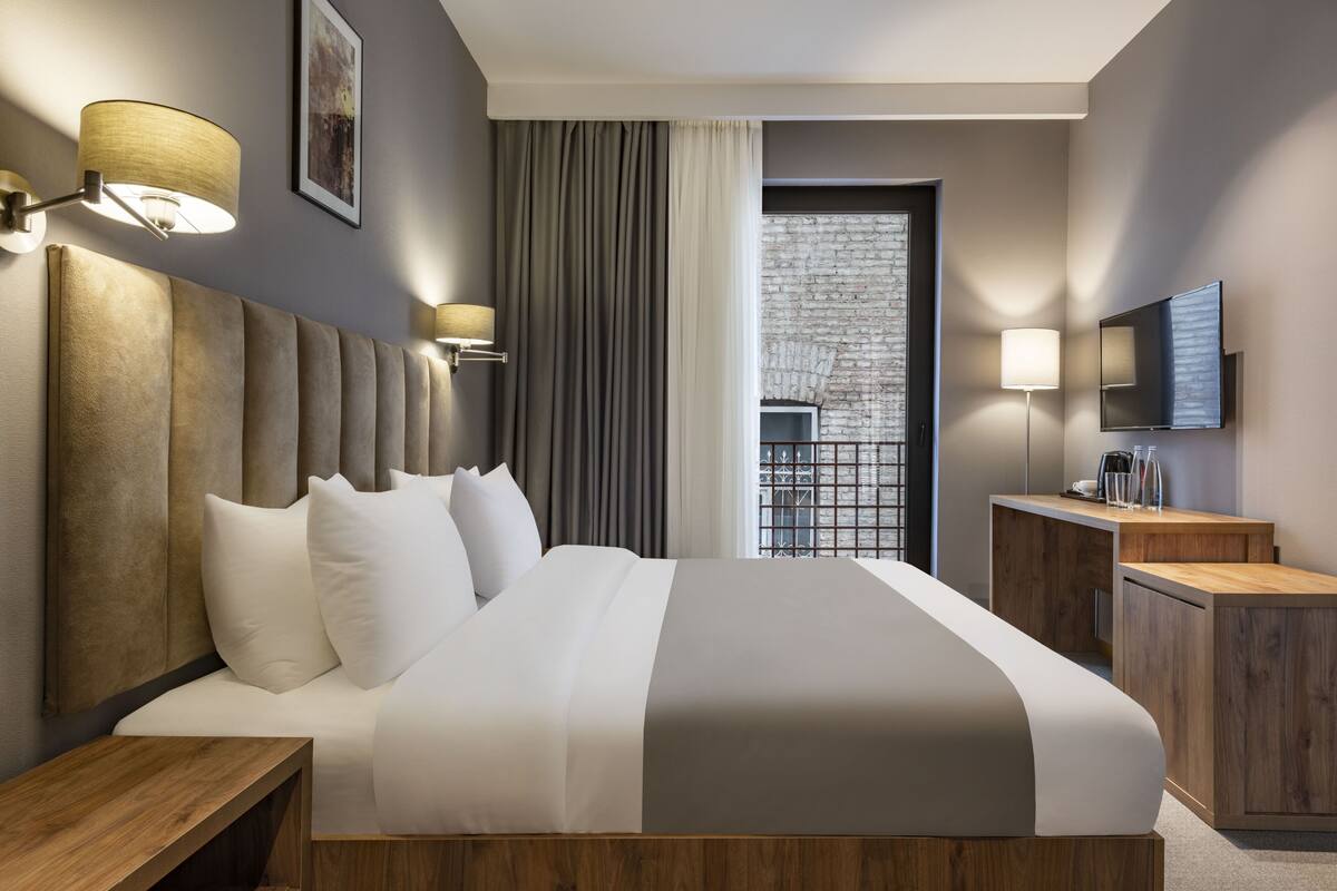 Habitación de hotel moderna con una cama grande, una mesita de noche de madera, un escritorio, un televisor de pantalla plana y vistas a un edificio de ladrillo a través de la ventana.