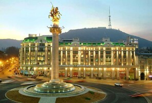 A hotel with a central fountain, multiple floors, and a view of a hill with a tower in the background. - BARNOVI BOUTIQUE HOTEL (Tbilisi)