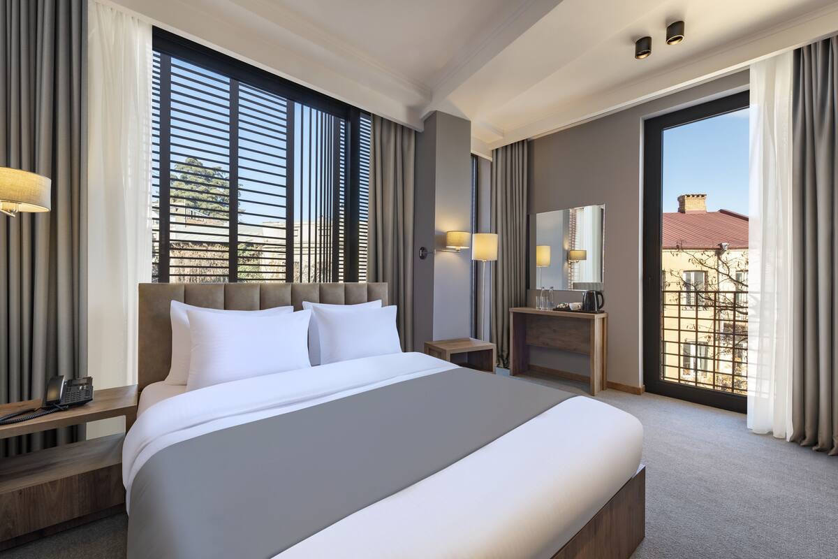 Habitación de hotel con una cama grande, mesitas de noche, un escritorio con lámpara, un espejo y vistas a la ciudad a través de amplios ventanales.