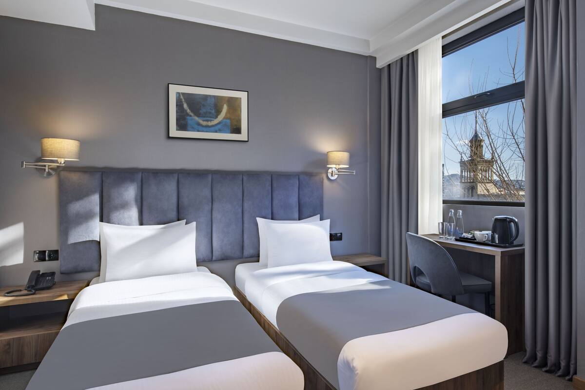 Habitación de hotel con dos camas, un escritorio, una silla, una lámpara, un cuadro y una ventana con cortinas.
