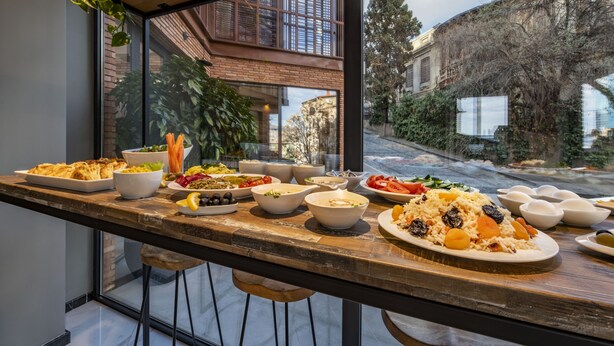A dining area with a long wooden table set with various dishes, large windows offering a view of a brick building and trees. - BARNOVI BOUTIQUE HOTEL (Tbilisi)
