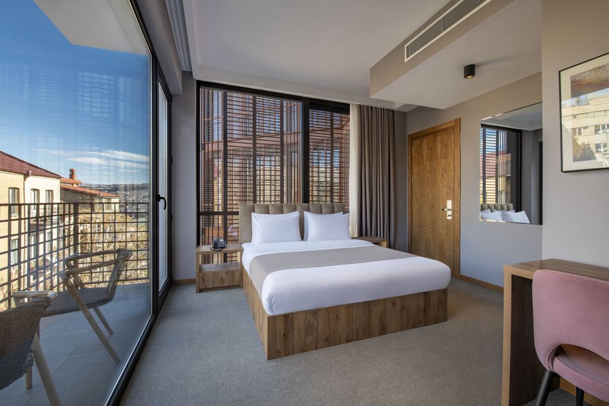 Una habitación de hotel moderna con una cama grande, un escritorio, una silla y un balcón con vistas a edificios.