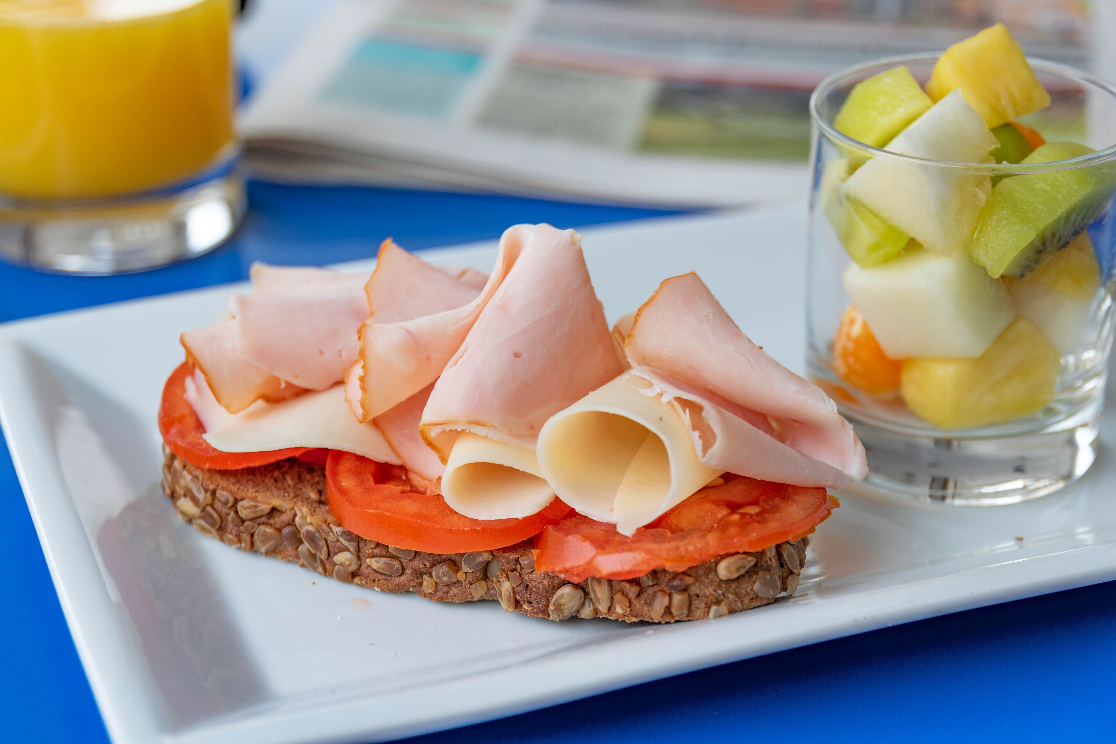 Un sándwich de jamón y queso sobre una rebanada de pan, acompañado de una ensalada de frutas y un vaso de jugo de naranja.