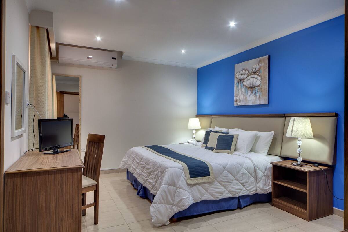 Habitación de hotel con cama, lámparas de noche, un escritorio con computadora y una pared azul con un cuadro floral.
