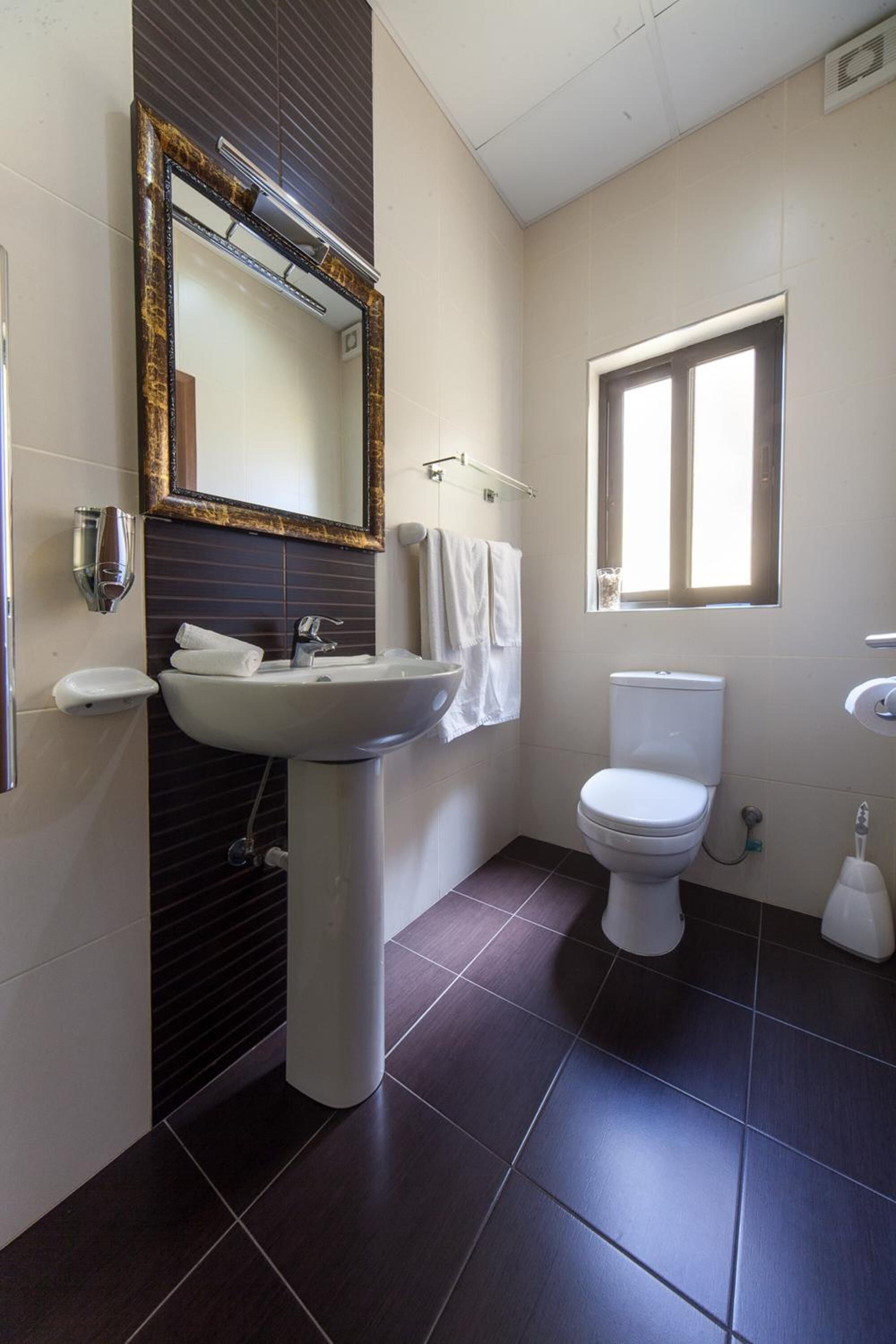 A bathroom with a pedestal sink, a toilet, a window, and a mirror with a decorative frame.