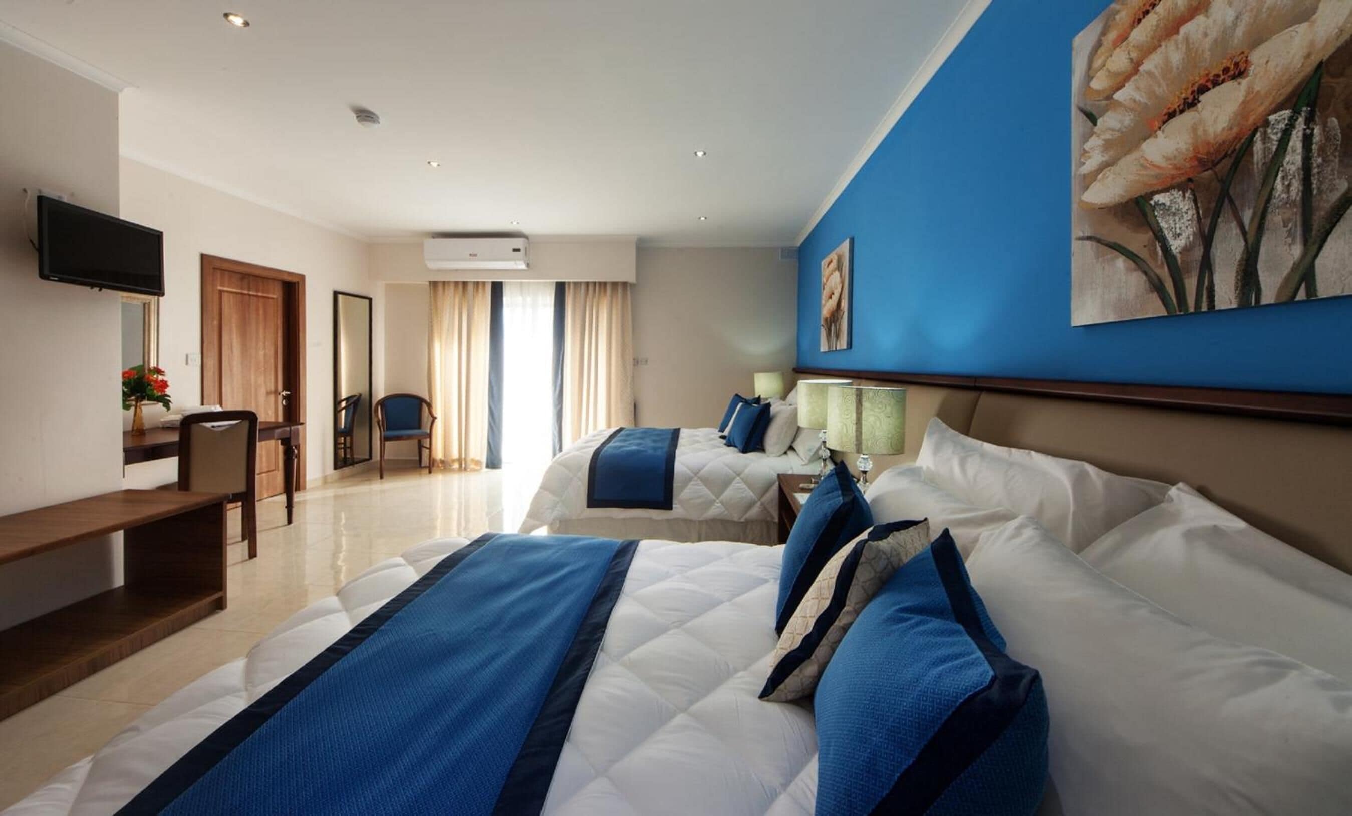 Habitación de hotel con dos camas, un escritorio, una silla, un televisor y un ventanal con cortinas.
