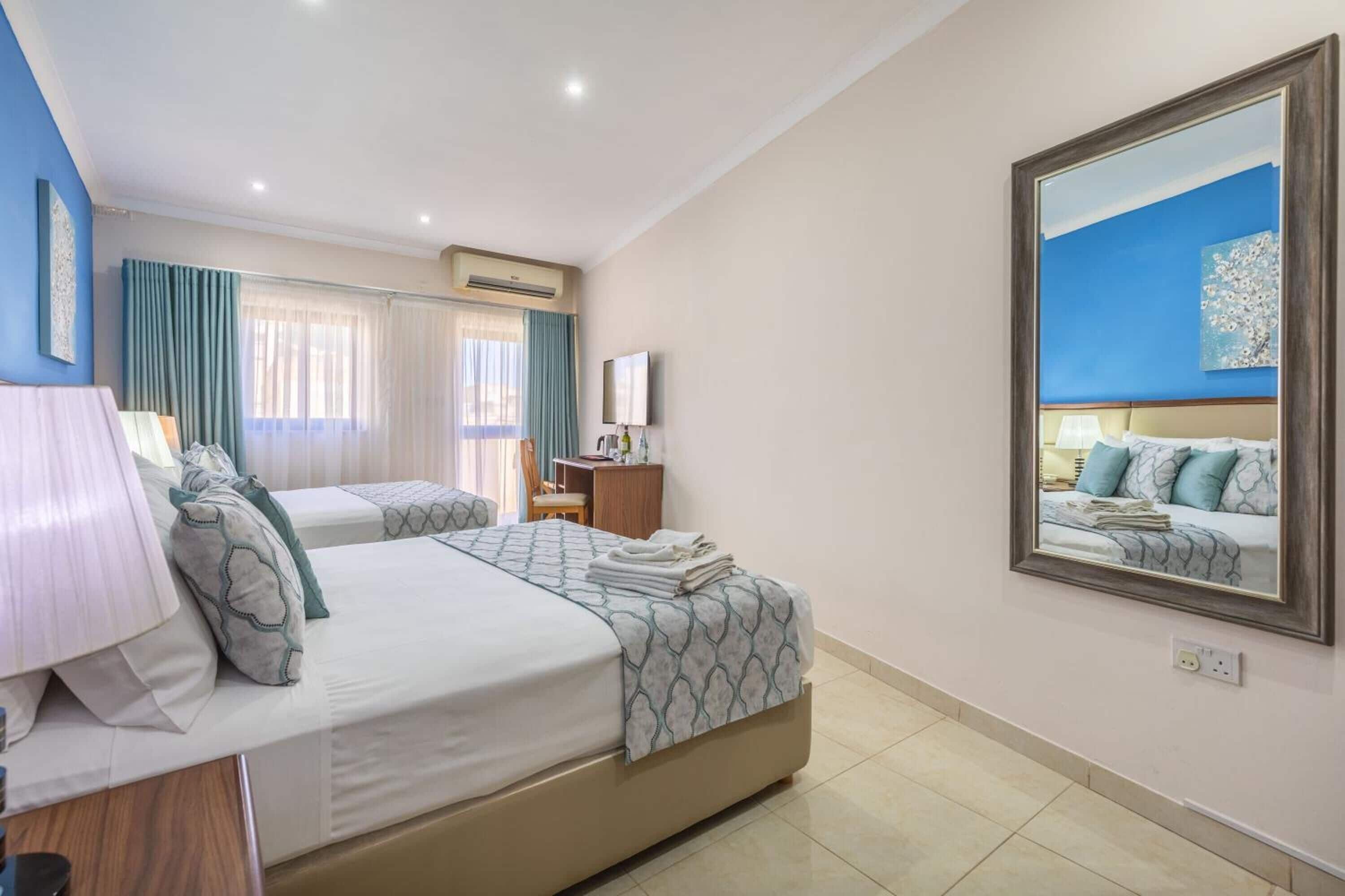 A hotel room with two beds, a desk, a chair, a mirror, and a window with curtains.