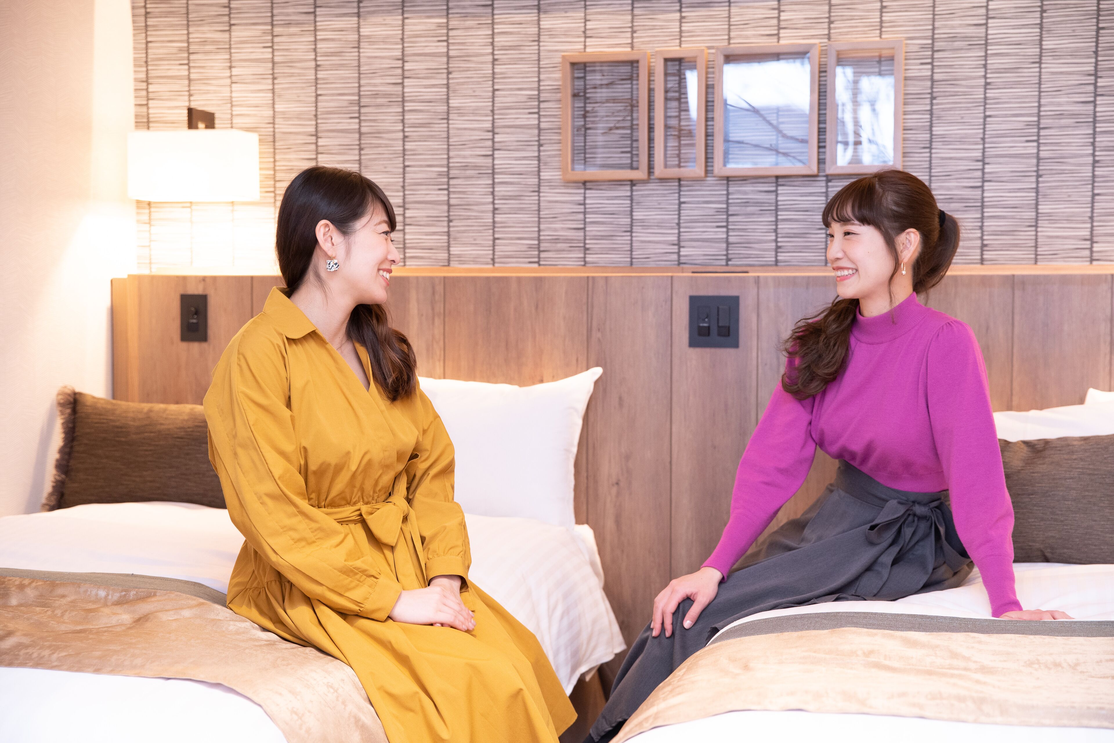Two women sitting on a bed in a hotel room with wooden headboard and framed pictures on the wall.
