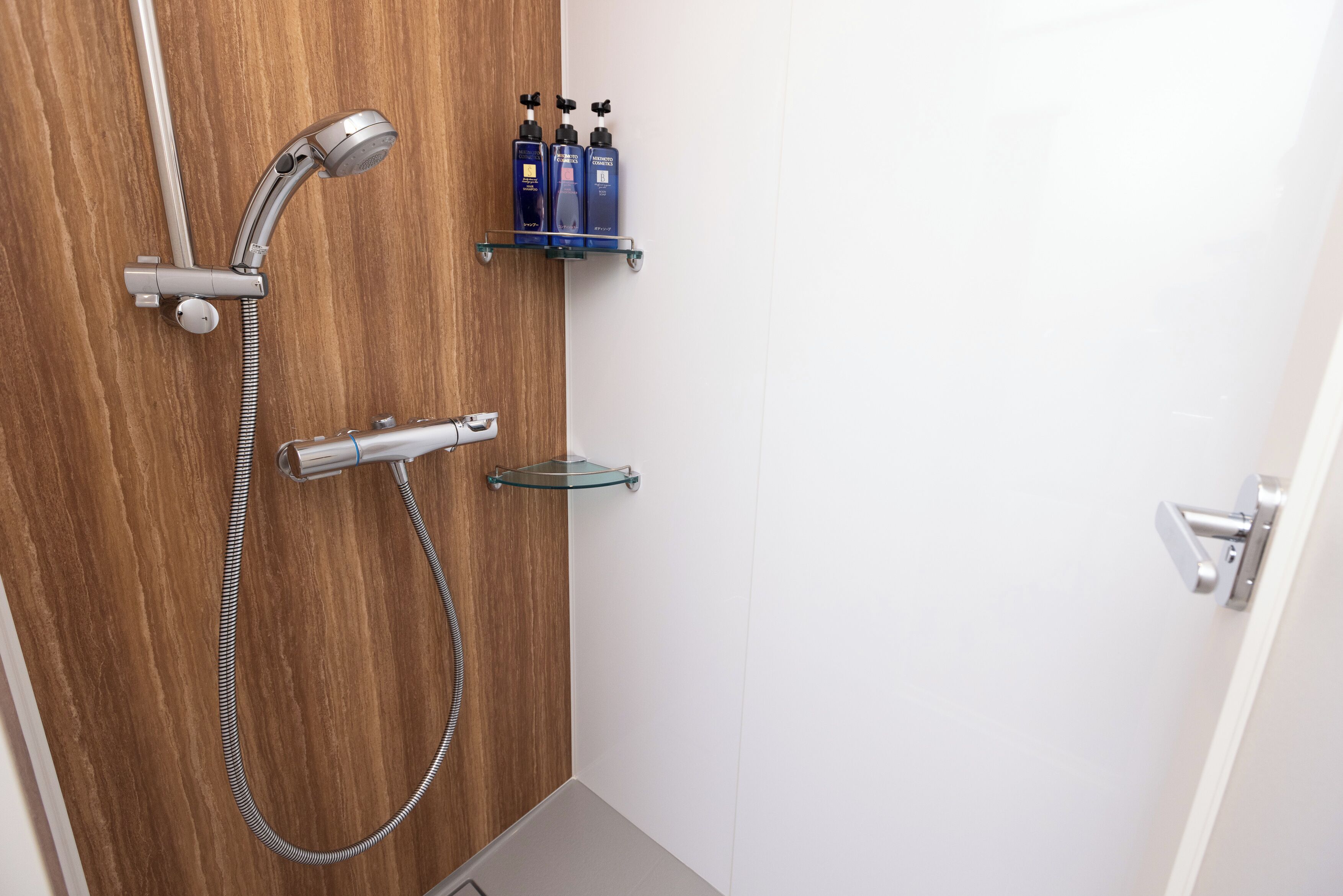 A shower with a handheld showerhead and a wall-mounted faucet, a glass shelf with two bottles, and a door handle.