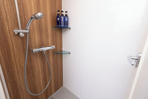 A shower with a handheld showerhead and a wall-mounted faucet, a glass shelf with two bottles, and a door handle.